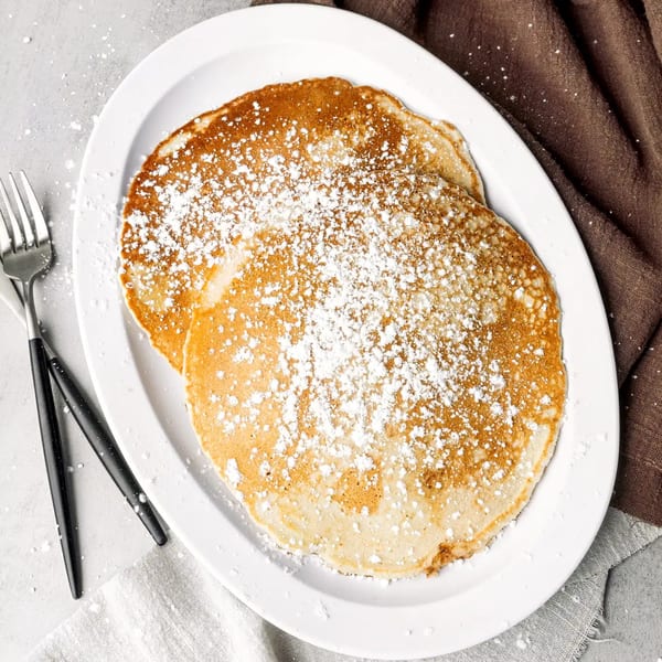 Two golden pancakes dusted with powdered sugar on a white oval plate with a fork.