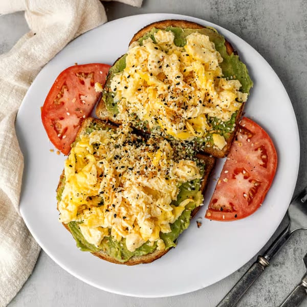 Two slices of avocado toast topped with scrambled eggs, everything bagel seasoning, and tomato slices.