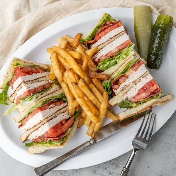 NY Deli club sandwich with turkey, bacon, lettuce, tomato, fries, and pickles on a white plate.