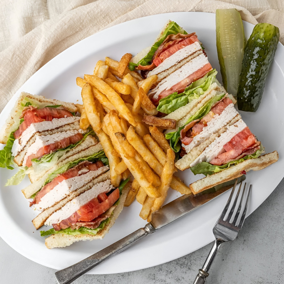 NY Deli club sandwich with turkey, bacon, lettuce, tomato, fries, and pickles on a white plate.