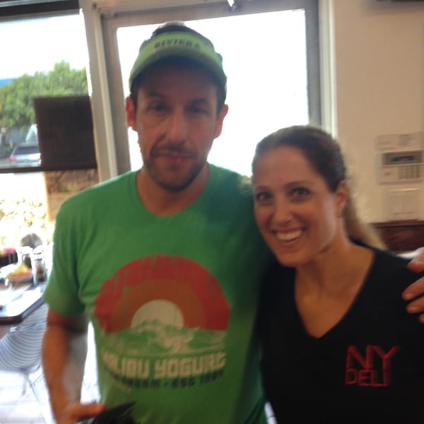 Adam Sandler and a smiling woman in an NY Deli shirt pose together inside the restaurant.