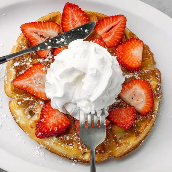 Golden waffle topped with fresh strawberries, whipped cream, and powdered sugar, with a fork and knife.