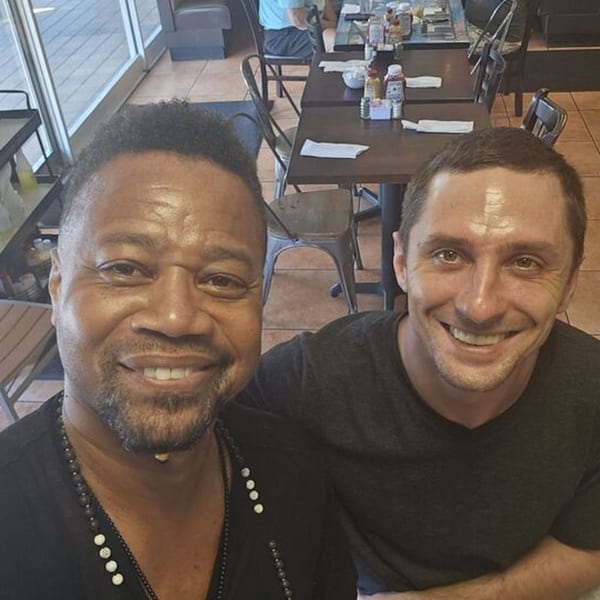 Two smiling men taking a selfie inside NY Deli, with restaurant tables and chairs in the background.
